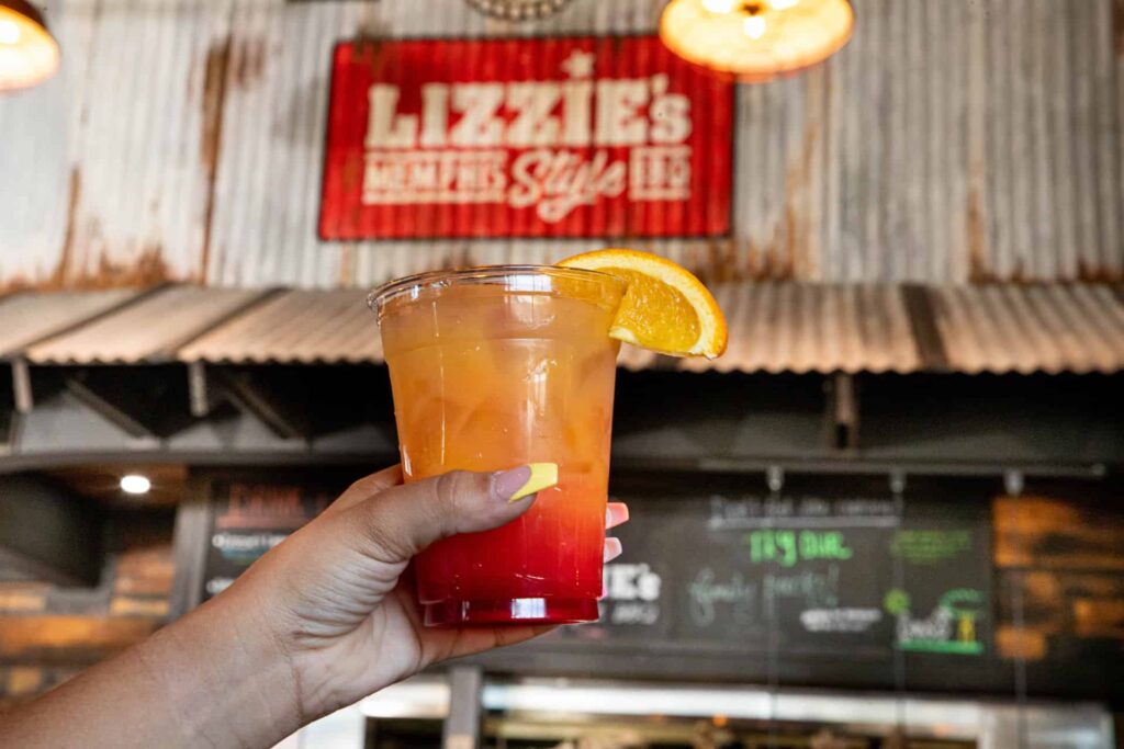 Hand holding a drink garnished with an orange in front of the Lizzie’s BBQ sign